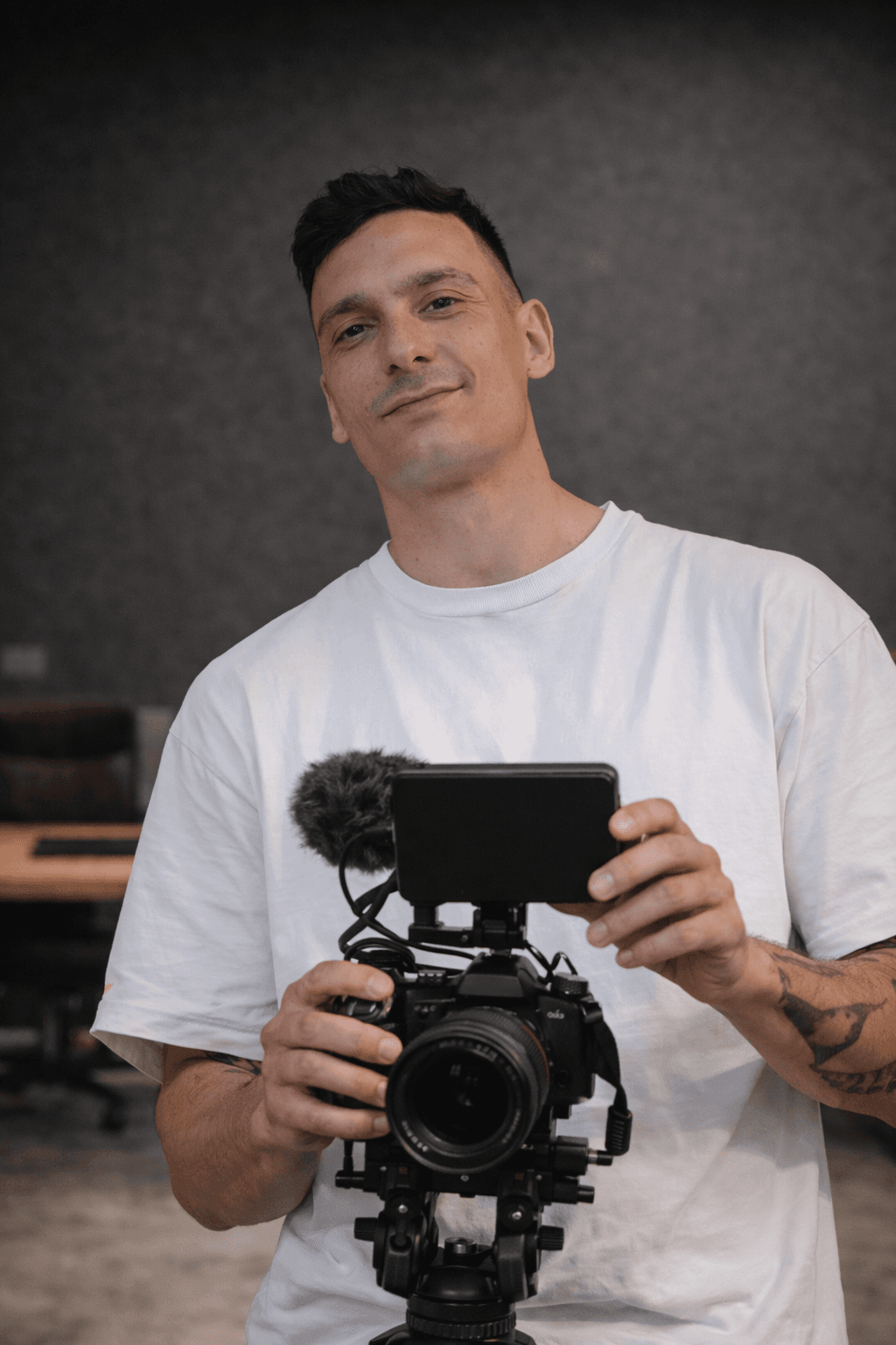 Uomo in maglietta bianca che tiene una videocamera professionale con monitor esterno e microfono.