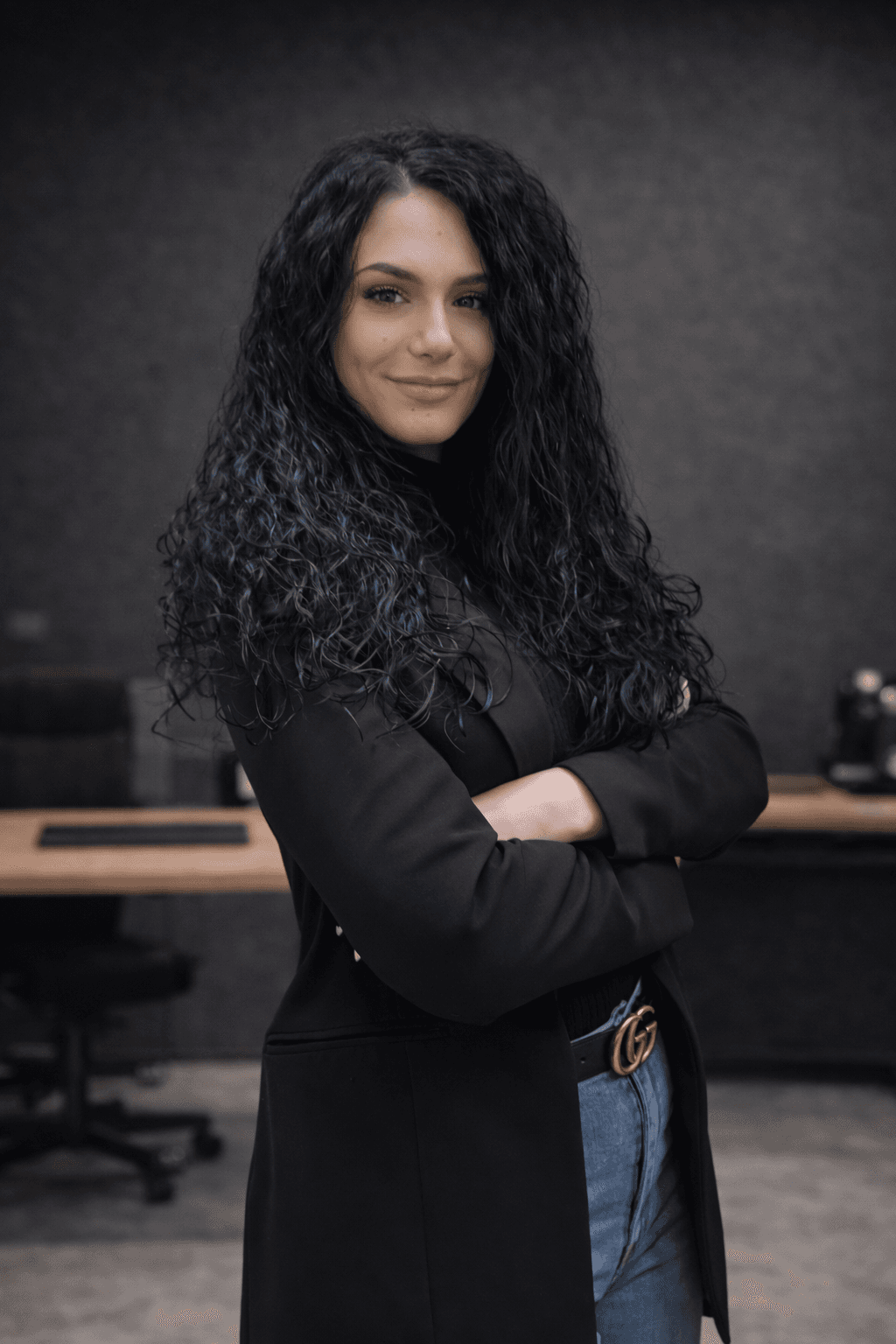 Donna con lunghi capelli ricci neri e blazer nero posa con braccia incrociate in ufficio.