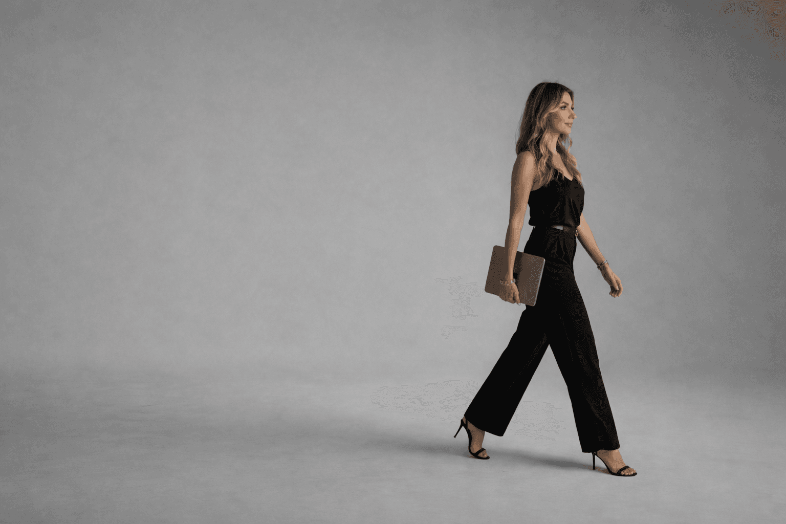 Donna bionda in tuta nera e tacchi cammina con un laptop su sfondo grigio.