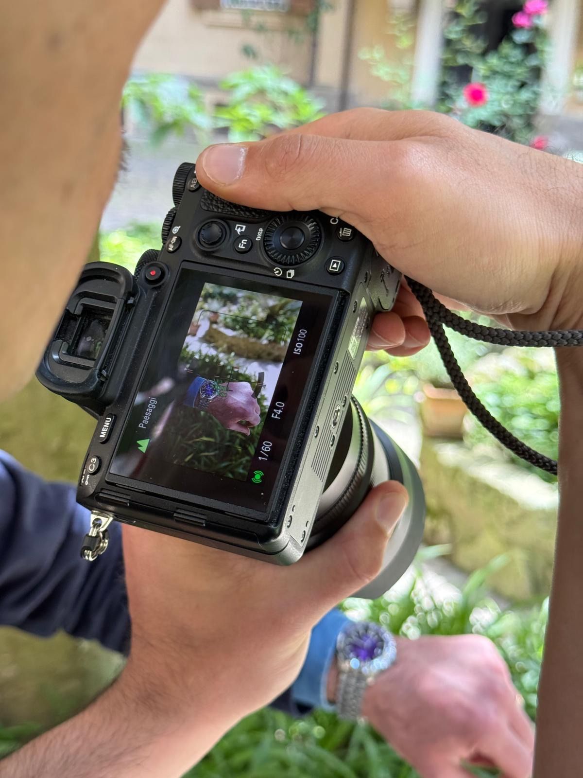 Mani che impugnano una fotocamera digitale inquadrando un orologio da polso in un giardino.