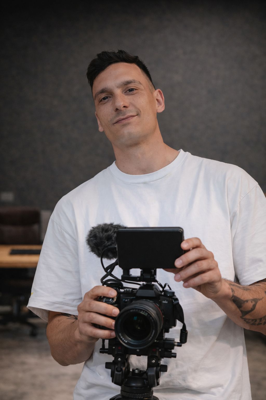 Uomo in maglietta bianca che tiene una videocamera professionale con monitor esterno e microfono.