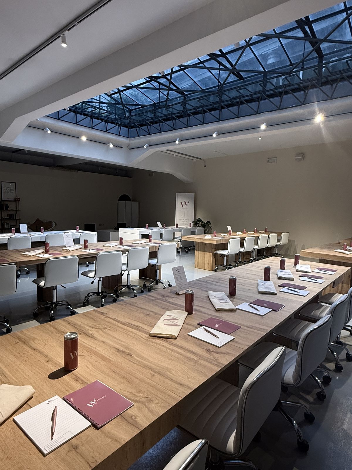 Moderna sala conferenze con tavoli in legno, sedie bianche e ampio lucernario in vetro.