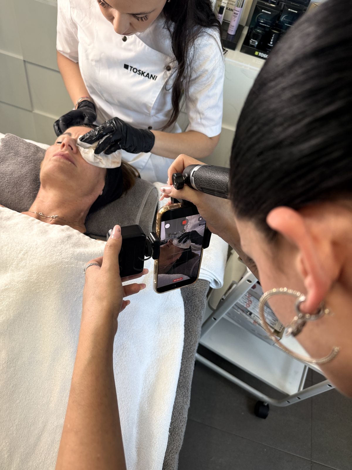 Estetista in guanti neri esegue un trattamento viso mentre una persona filma con uno smartphone.