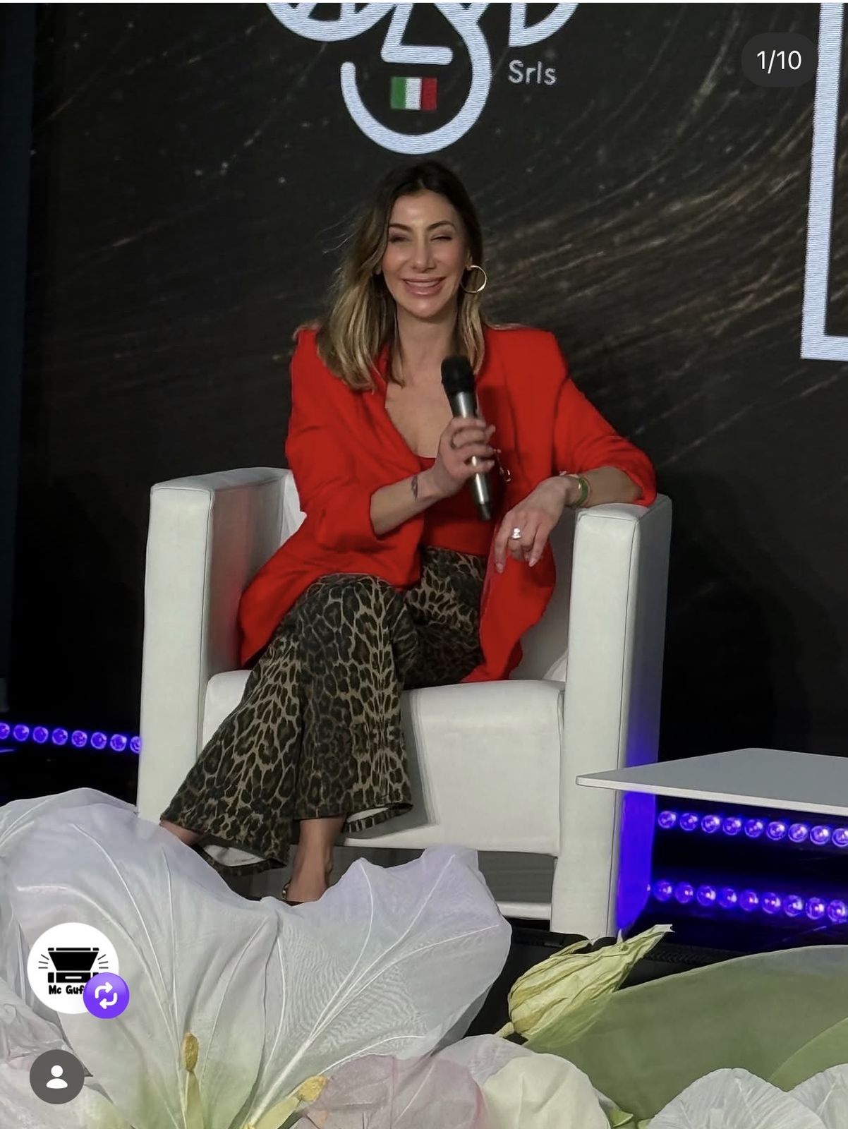 Donna sorridente in blazer rosso e pantaloni leopardati seduta su una poltrona bianca con microfono.